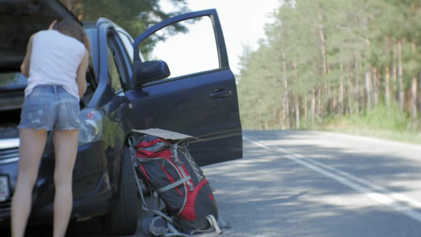 Young beautiful woman hitchhiking standing on the road with a backpack on a table with an inscription SOUTH near a broken car 4k