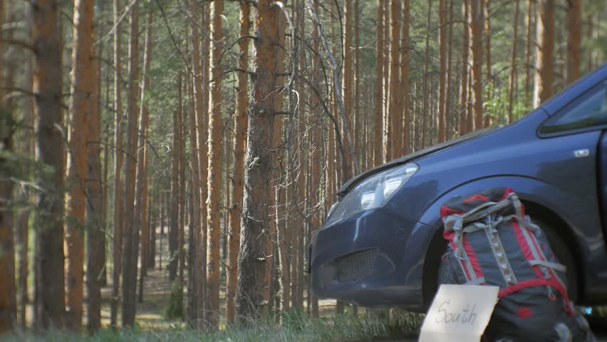 Young beautiful woman hitchhiking standing on the road with a backpack on a table with an inscription SOUTH near a broken car 4k