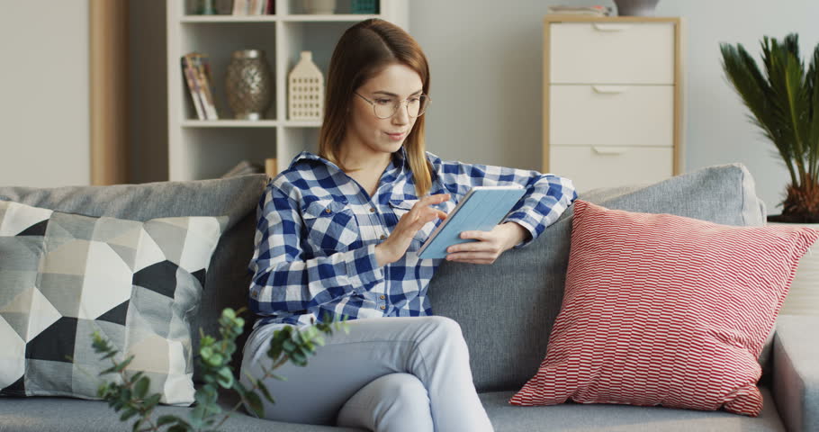 Beautiful young woman sitting on the couch at home, watching something on the tablet device and laughing. Indoor