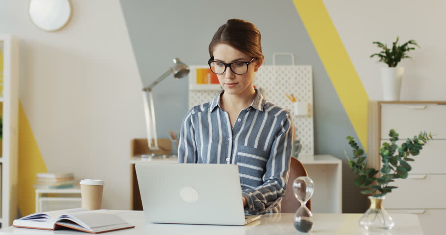 Portrait of the Caucasian young pretty woman in the striped blouse and glasses working at the laptop computer in the office and then smiling to the camera. Inside