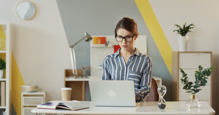 Young succesful businesswoman in the glasses working in the office room at the laptop computer and thinking while typing. Indoors