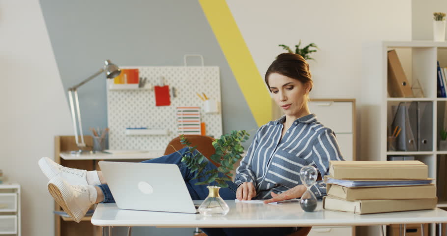 Young beautiful woman sitting at the office table, holding her legs on it, making a paper plane and throwing it away. Indoor