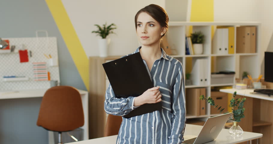Portrait shot of the young pretty woman with a black folder in hands standing and posing in the modern office room. Indoors