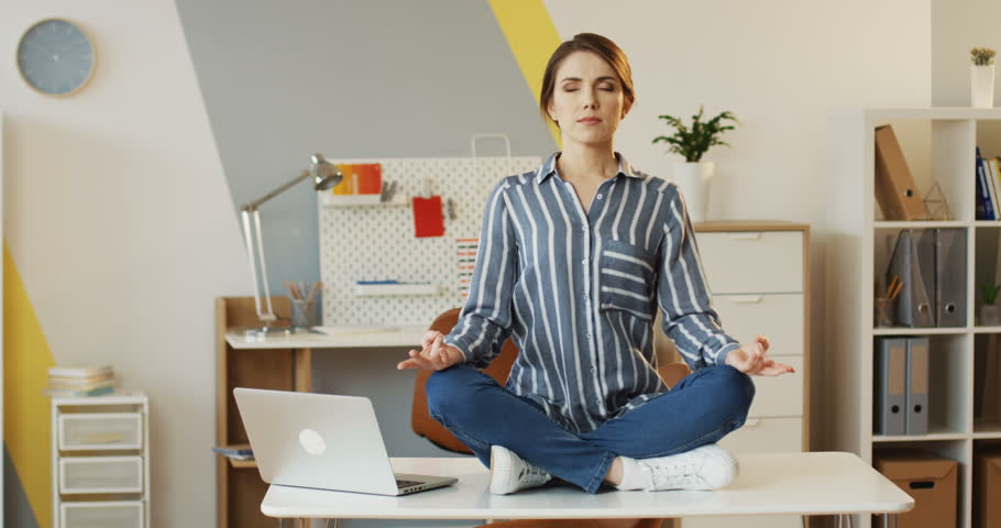 Charming young woman in the striped shirt sitting on the office table near laptop and meditating in the lotos pose. Indoors