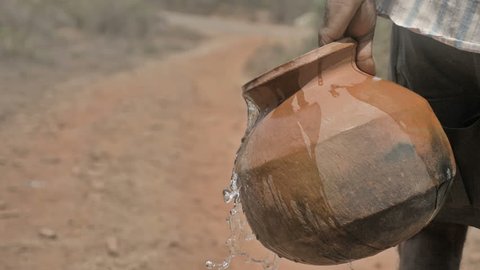 Water spilling out of a earthen pot of a farmer walking on barren terrain in rural road. A poor man carrying clay pot filled with water while some water is spilling out in drought affected region
