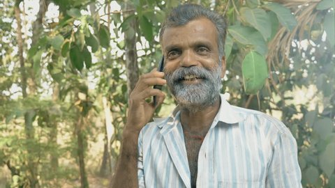 A middle aged bearded cheerful man talking on a cellphone or smartphone in the farm surrounded by green trees. An experienced male farmer speaking on mobile phone in the fertile agricultural land 