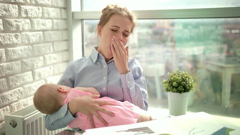 Tired woman yawn with sleeping child on hands. Tired mother yawning with baby and checking smartphone. Sleepy mom with kid at workplace. Yawning woman with baby at office workplace near window