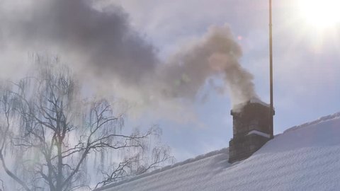 Smoking chimney of a wood fire heated house in a cold sunny winter day, snow on a roof