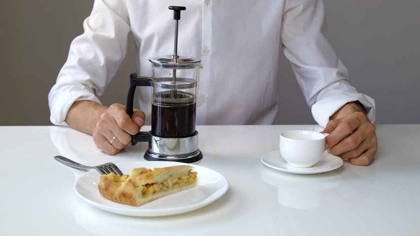 Poring black coffee out of french press brewer and drinking it. Young male person in white business shirt having lunch or breakfast in minimalistic background