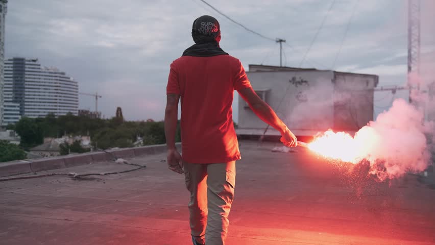 Young man in balaclava with red burning signal flare on the roof with graffiti background, slow motion
