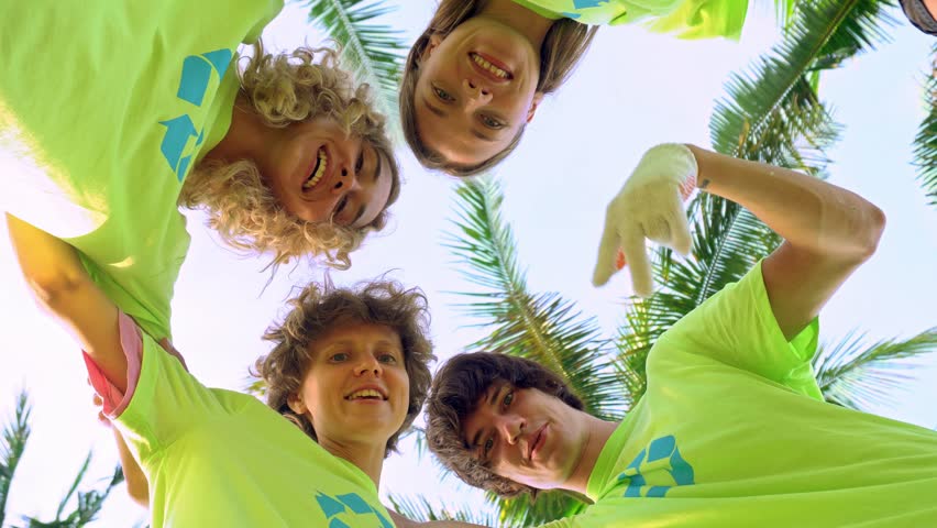Four young volunteers in green t-shirts with a picture of recycle forming huddles under palm trees on the shore of an ocean beach, after cleaning the beach from plastic debris. Volunteering and Royalty-Free Stock Footage #1011578501