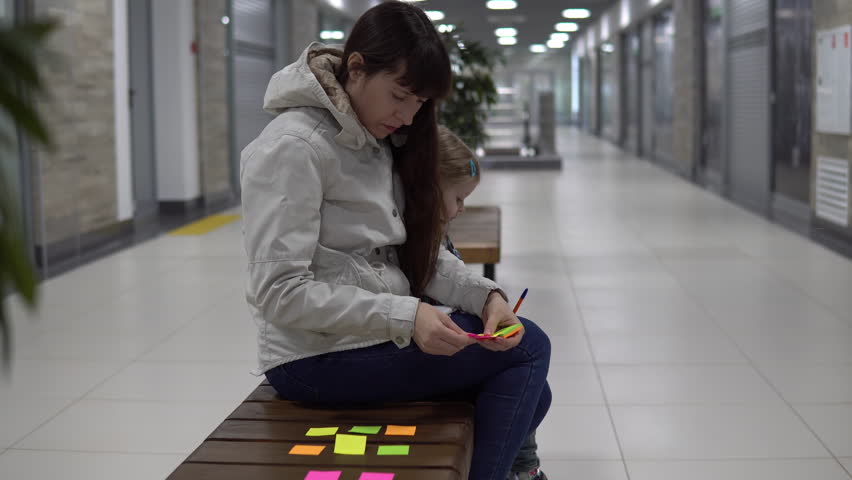 A young woman checks her notes with a shopping list sitting on a bench in the mall with her little daughter.