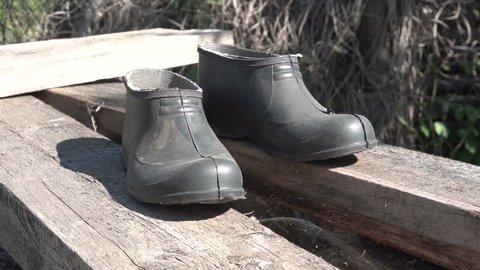Rubber galoshes stand on wooden boards. Pair of shoes. Black. Summer time. Bright day. Green leaves on the background. Country life. Rustic atmosphere.