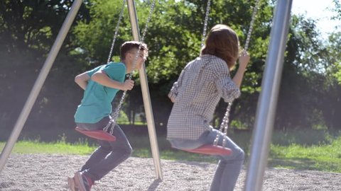 Happy two children ride on a swing at summer park. Cute teen boy and girl swings at playground outdoors.