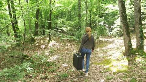 girl, walks through the woods with a heavy black suitcase. 4k, slow-motion shooting, steadicam shot.