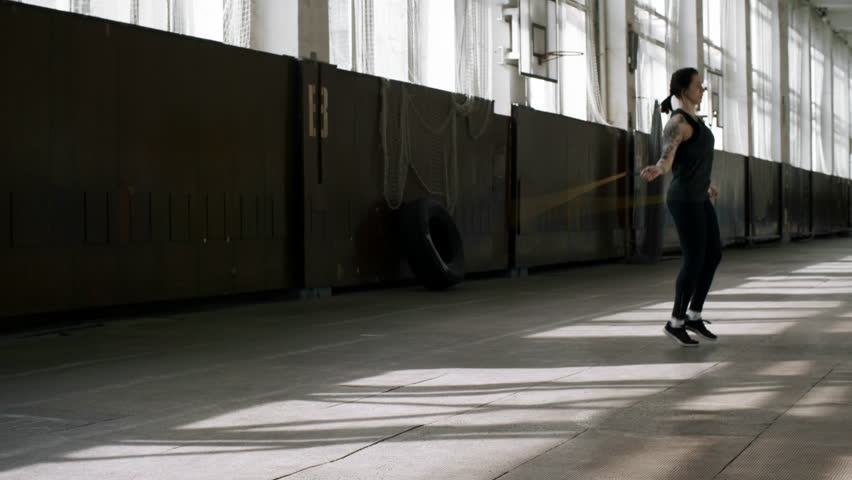 Dolly shot of sweaty sportswoman with tattoos breathing hard and jumping rope n empty gymnasium