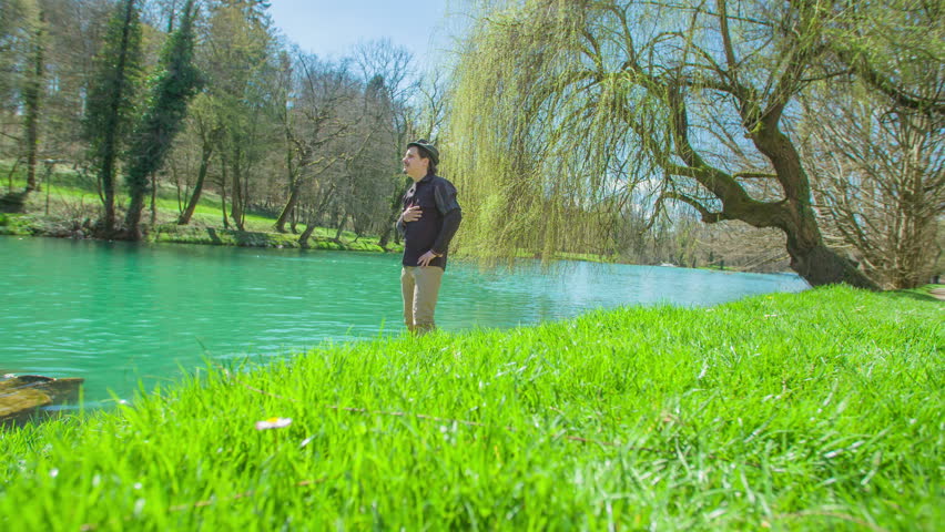 A man is standing by the river and he is looking into the water. His hand is on his chest.