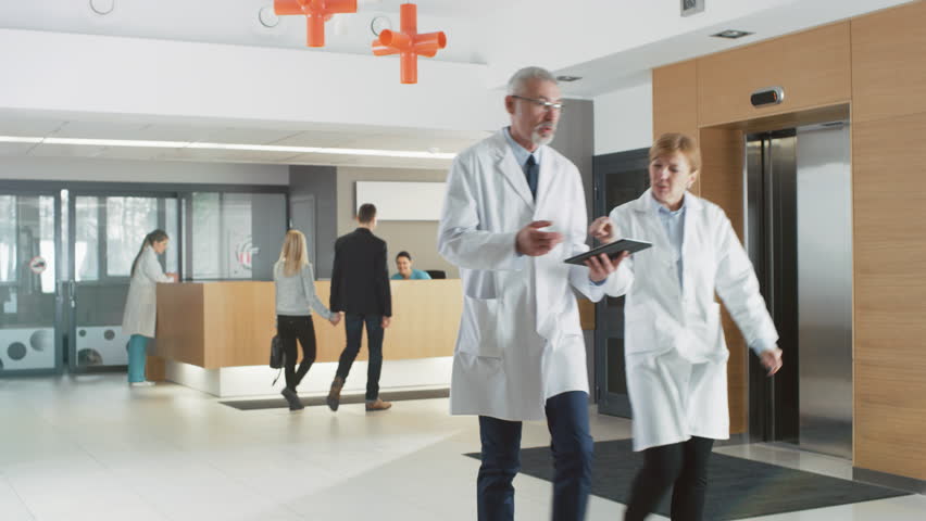 In the Hospital Young Couple Walks to Reception. Medical Personnel, Doctors, Nurses, Assistants and Patients in the Busy Lobby. New, Modern Medical Facility. Shot on RED EPIC-W 8K Helium Cinema Camera