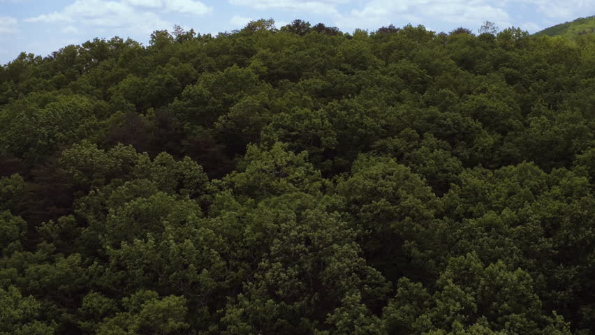 Flying over crest of hill in southern Blue Ridge Mountains in north Georgia with clouds and sunlight, lake or river in distance, May 2018 