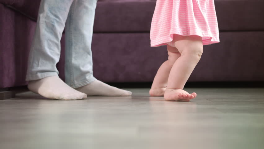 Little baby feet walking on floor with parent support. Close up of mother learning child walking. Kid feet steps at home. Little child learn to walking. Baby steps with mom. Toddler doing first step