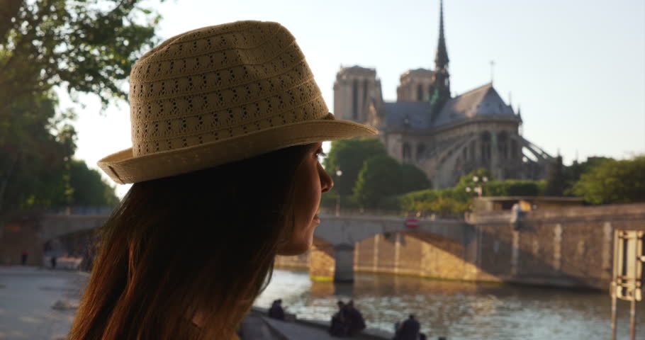 Side view of tourist woman in fedora looking out at the Seine. Pensive female near Notre Dame Cathedral sightseeing and exploring Paris. 4k