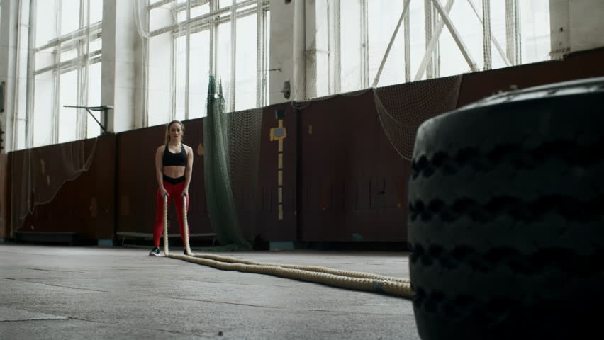 PAN of strong young sportswoman in fitness top and legging breathing hard and battling rope in empty gymnasium during conditioning workout routine