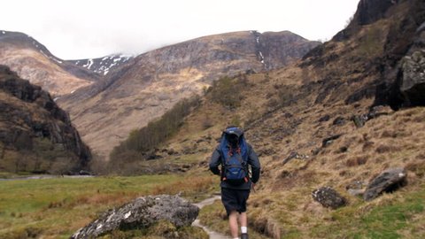 4K Young traveller hiking through the Scottish Highlands. Man nature path walking and back pack trekking through rural valley and mountains. A natural rocky landscape of a British tourist exploring 
