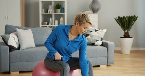Caucasian middle aged woman in sporty style sitting on the fitness ball and swinging her hands with dumbbells at home. Indoors