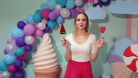 A young girl in bright clothes with lollipops in her hands in a studio with balloons, marshmallows and ice cream, she can not choose one lollipop of two. Barbie style. Candy girl.