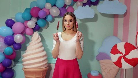 A cheerful girl with big lollipops in her hands is having fun in a bright room with balloons and a large plastic ice cream, she is dressed like a barbie. Barbie girl. Candy girl.
