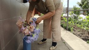 old woman with short curly hair and excess weight puts flowers in the water in the blue watering pad. medium plan. Then grandma cut off the dry inflorescences.