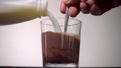 Slow motion close shot of a man’s hand stirring milk from a plastic carton into a glass of chocolate powder mix, to make a chocolate milk drink.