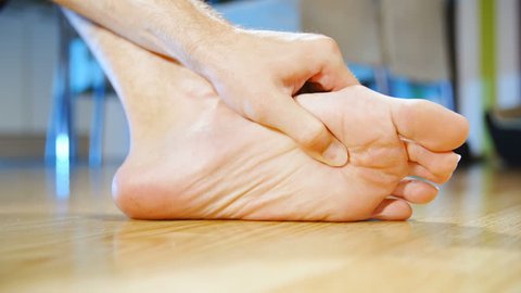 Massaging painful foot by hand slow motion 4K. Low angle long shot of person's foot in focus while massaging with a hand at home. Background dining room out of focus.