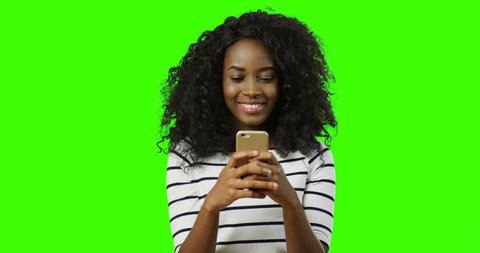 Pretty cheerful african american young woman with curly hair chatting with somebody on the smartphone while typing and smiling on the green screen background. Chroma key.