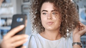 Video portrait like curly beautiful young woman smiles to the camera and makes selfie with her phone in different poses, kinky girl makes photos to the social networks, photos after making hairstyle
