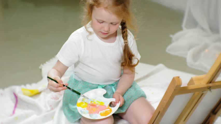Beautiful red-haired girl sits on the floor in the studio in front of the easel and draws a brush and colored paints 4k