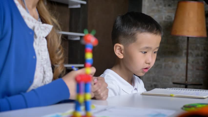 Asian little kid taking falled pen from floor and studing with mother, sitting behind table, book shelves and brick wall background
