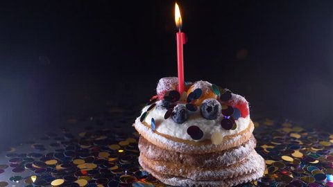 holiday, celebration and party concept - birthday cupcake with one burning candles over black background, confetti. Confetti for a party. super slow motion