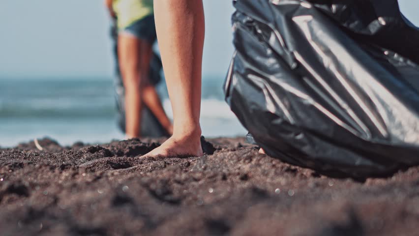 Group of volunteers cleaning up beach. The volunteer raises and throws a plastic bottle into the bag. Volunteering and recycling concept. Environmental awareness concept copy space | Shutterstock HD Video #1011722213