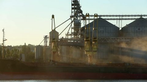 Panorama of grain terminal at seaport on sunny day. Cereals bulk transshipment to vessel loading grain crops on bulk ship from large elevators at the berth. Transportation of agricultural products.