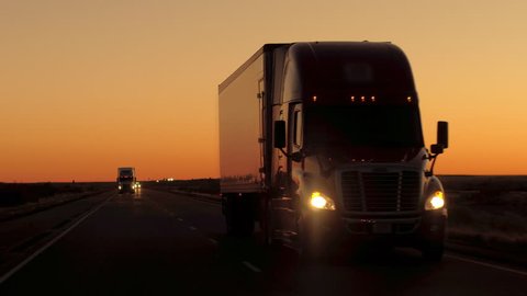 CLOSE UP: Front view of semi trailer trucks with freight container transporting goods driving along the scenic country highway after the sunset. Lorries hauling cargo on across vast grassy plains