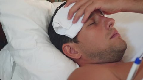 A sick man lies in a bed with a thermometer and a towel on his head, from a strong headache with pain. slow motion, 1920x1080, full hd