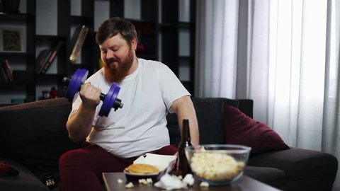 Funny fat guy eating fast food, drink beer and doing dumbbell training. He is sitting at home on the couch in front of the TV. Concept of malnutrition, sport, obesity