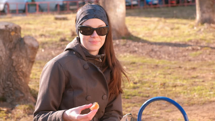 Young brunette woman in sunglasses eats tangerine sitting on the bench in park.