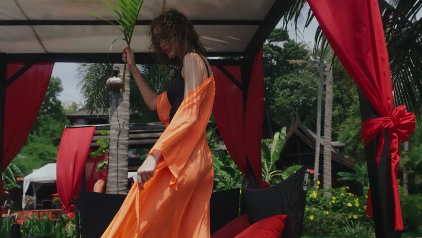 Young curly girl fashion model posing in an orange pareo under palm trees next to sun beds in a warm tropical country on a hot sunny day