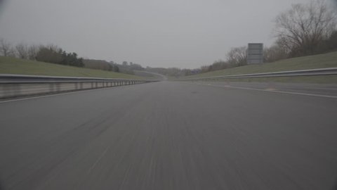 Low level shot of road textures, pulling backwards, motorway style road on a circuit track. Grey skies with grass verges on the side. 