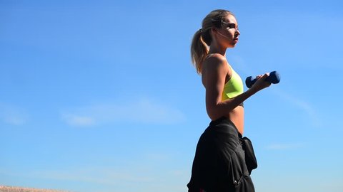 Beautiful, athletic young blond woman doing exercises with weights, dumbbells, lunges, squats. City embankment, blue sky at the background, summer sunny morning. 