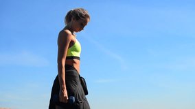 Beautiful, athletic young blond woman doing exercises with weights, dumbbells, lunges, squats. City embankment, blue sky at the background, summer sunny morning. Fitness in the city healhy lifestyle