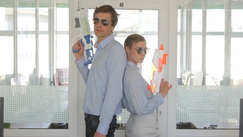crop close up view of young man and woman standing in sunglasses backs to each other with toy pistols in office