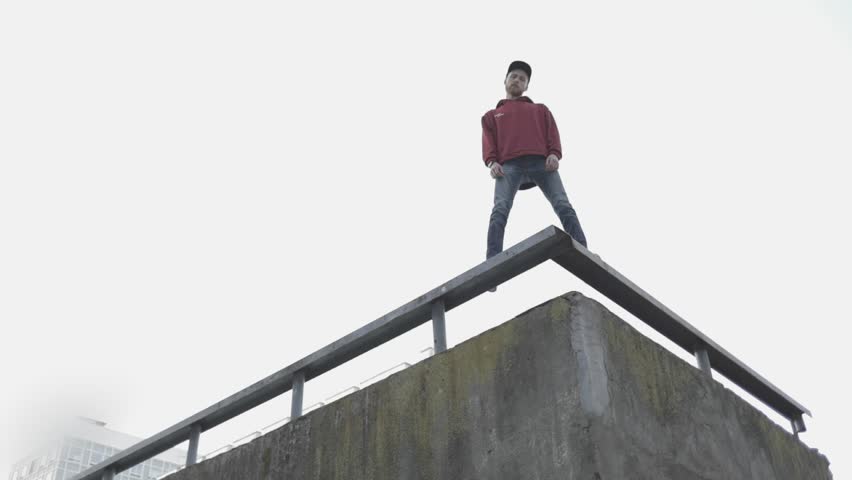 The view from below is a handsome strong guy standing on the edge of a building roof. A man in a red sweatshirt looking down at the camera.